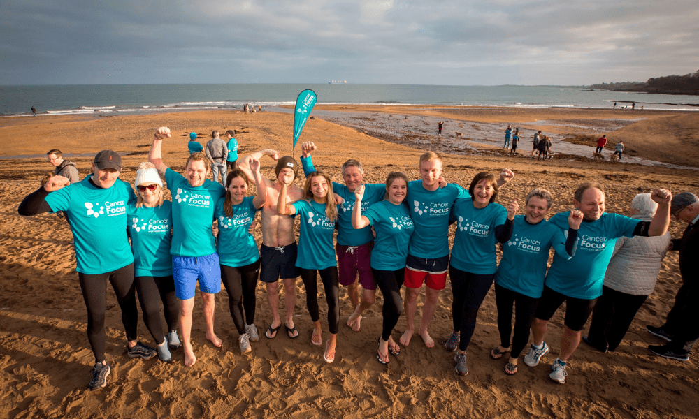 Cancer Focus NI Dare to Dip event. Group of swimmers after entering the chilly sea wearing Cancer Focus NI t-shirts. Background is of a beach. 