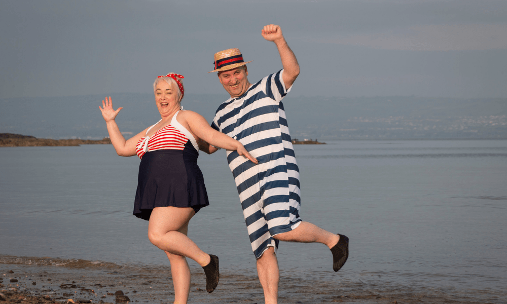 A man and a lady in fancy dress out fits, with one foot in the air and holding their hands up. The lady on the left is in a swimming costume with a red and white top and black skirt bottom, she had short blonde hair with a red and white bandana. The man has a straw hat with a red and black ribbon across it and his he wears a knee length navy and white striped dress. The background is the sea at Crawfordsburn beach.
