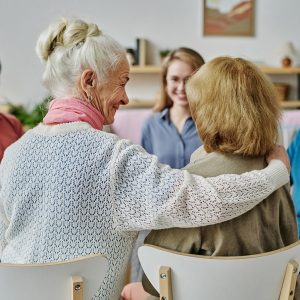 Rear view of senior woman supporting her friend while they sitting at psychotherapy session with other older people